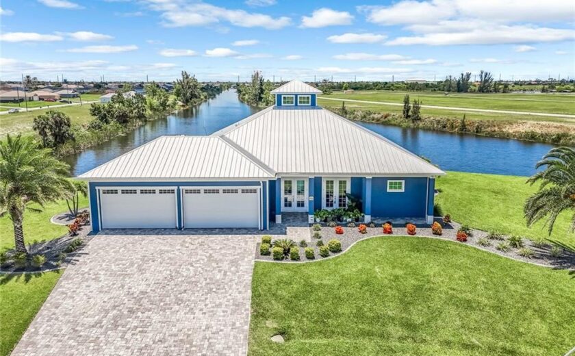 Lauren Homes coastal custom home with a silver metal roof, cupola, blue exterior, three-car garage and paver driveway on a Cape Coral canal